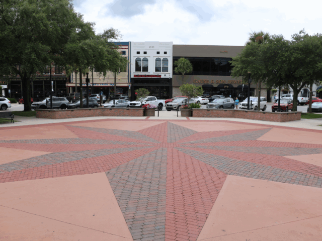 A wide, open town square with a red and gray brick star pattern in the center, surrounded by low brick walls and benches. In the background, cars are parked along the street in front of several storefronts. Trees line the sidewalk, providing shade, and flower planters add color to the scene.
