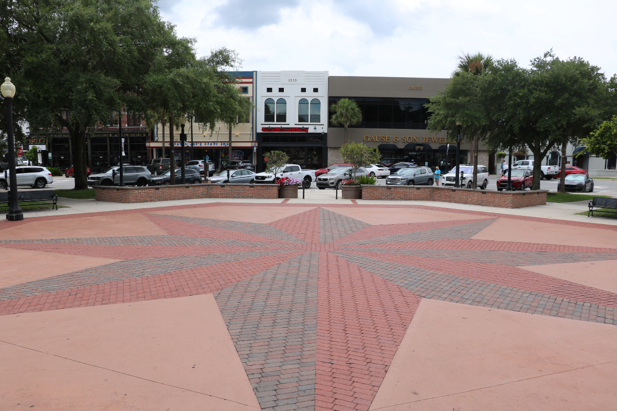 A wide, open town square with a red and gray brick star pattern in the center, surrounded by low brick walls and benches. In the background, cars are parked along the street in front of several storefronts. Trees line the sidewalk, providing shade, and flower planters add color to the scene.