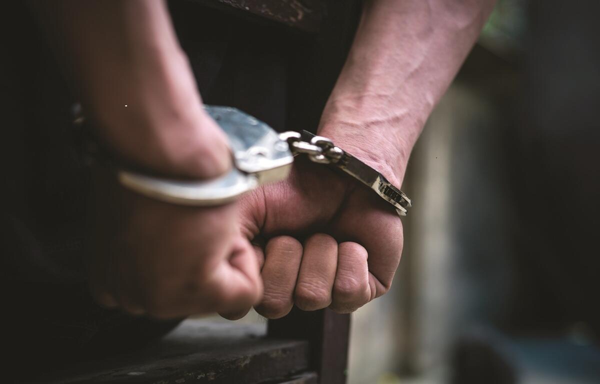 An individual standing in a blurred background with their arms behind their back, handcuffed.