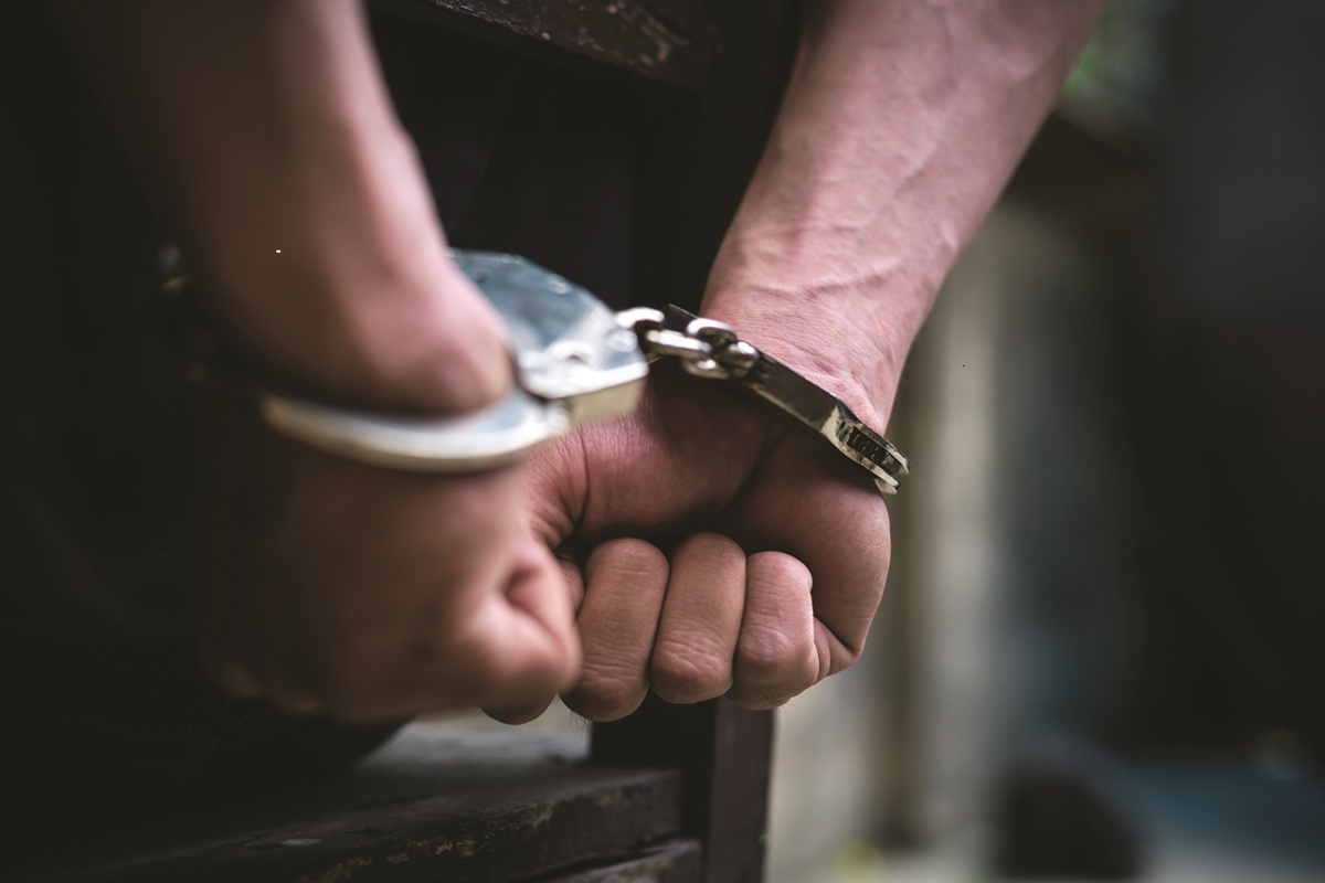 An individual standing in a blurred background with their arms behind their back, handcuffed.