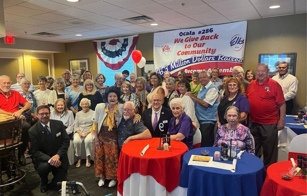 A group of men and women standing and sitting in front of a wall with a patriotic banner and a colorful poster with letters and numbers against the back wall. There are tables with red, white and blue tablecloths to the right.