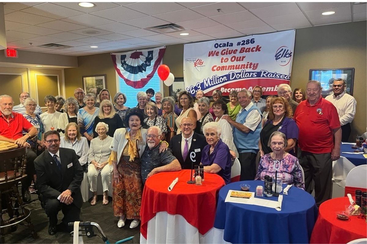 A group of men and women standing and sitting in front of a wall with a patriotic banner and a colorful poster with letters and numbers against the back wall. There are tables with red, white and blue tablecloths to the right.