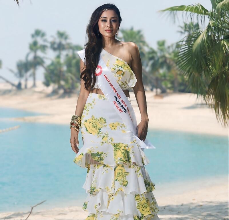 Nilam Patel wearing a floral dress and sash, standing on a sandy beach with palm trees and turquoise water in the background.