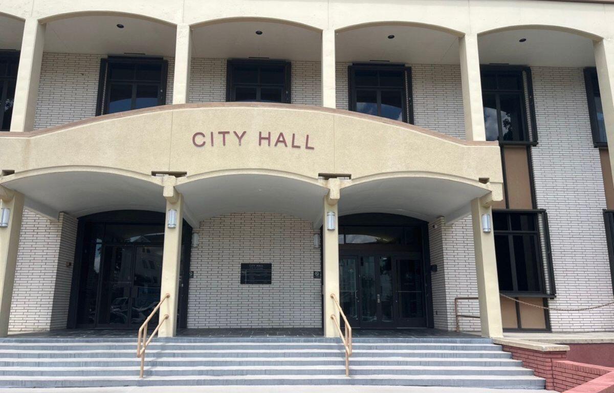 A building with a white and beige facade, with the beige pat having lettering, there is a plaque in the far background, and columns on both the first and second structure, with gray steps and guard rails for the step in the foreground.