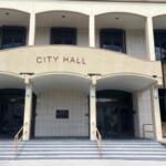 A building with a white and beige facade, with the beige pat having lettering, there is a plaque in the far background, and columns on both the first and second structure, with gray steps and guard rails for the step in the foreground.