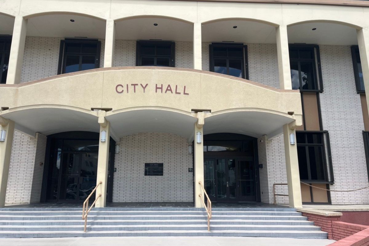 A building with a white and beige facade, with the beige pat having lettering, there is a plaque in the far background, and columns on both the first and second structure, with gray steps and guard rails for the step in the foreground.