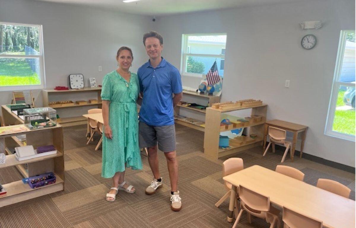 To the left in the background, bookcases with books and other items, along the far wall items on bookcases, tiny chairs and desks, an American flag by the window, a couple in the middle of the room standing on a carpeted floor. to the right in the foreground, tiny desks and chairs.