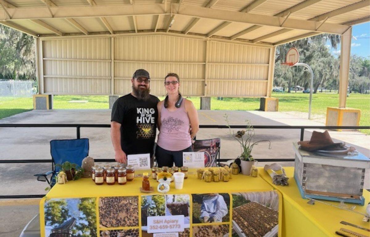 A man in a baseball hat stands next to a woman, with a table that has a yellow tablecloth, with a poster on the front of the table. There are a variety of items on the table. in the background there's the covered roof, pipe rail metal fence and a concrete floor.