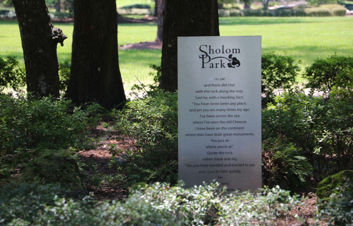 A sign that reads "Sholom Park" stands in the middle of bushes with tall trees in the background. An open green field is visible in the distance.