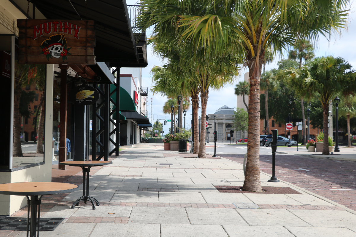 White-bricked sidewalk lined with palm trees and a red-brick road on the right. On the left are two circular peach-colored tables, Mutiny Bar Ocala, Ivy on the Square Boutique and other shops and restaurants in the distance on the left.