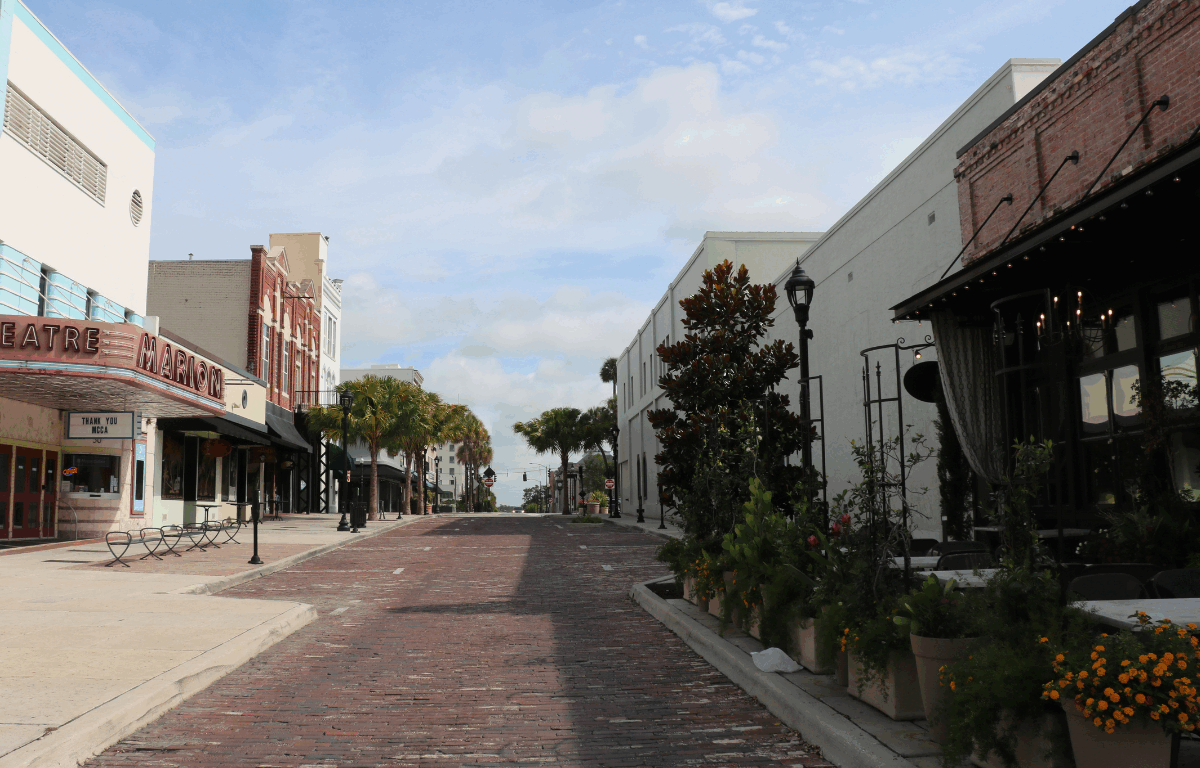 Brick paved street that begins narrow and widens further out with the Marion Theatre, The Tipsy Skipper, Mutiny Bar Ocala and The Lodge lining a white sidewalk on the left side. On the right side of the road lies Ivy on the Square with plants lining the sidewalk nearest the road and a white, vacant building. Various parallel parking spots line the brick paved road.