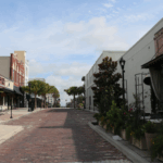 Brick paved street that begins narrow and widens further out with the Marion Theatre, The Tipsy Skipper, Mutiny Bar Ocala and The Lodge lining a white sidewalk on the left side. On the right side of the road lies Ivy on the Square with plants lining the sidewalk nearest the road and a white, vacant building. Various parallel parking spots line the brick paved road.