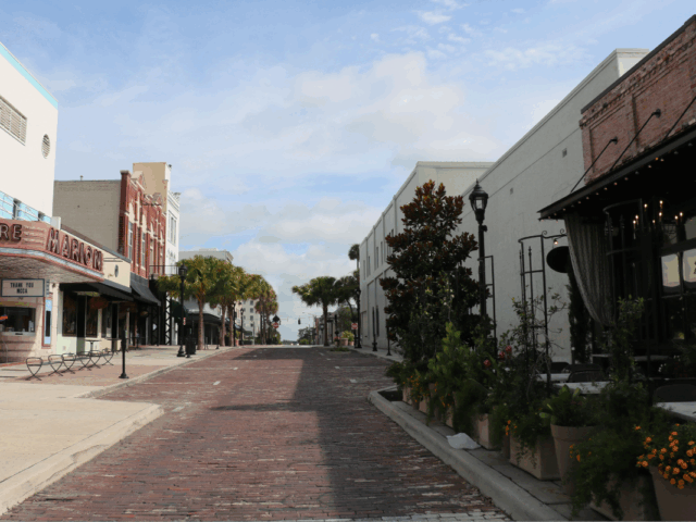 Brick paved street that begins narrow and widens further out with the Marion Theatre, The Tipsy Skipper, Mutiny Bar Ocala and The Lodge lining a white sidewalk on the left side. On the right side of the road lies Ivy on the Square with plants lining the sidewalk nearest the road and a white, vacant building. Various parallel parking spots line the brick paved road.