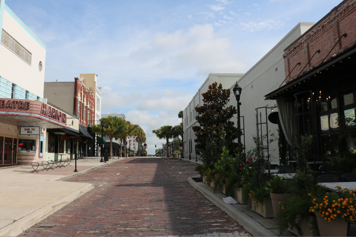 Brick paved street that begins narrow and widens further out with the Marion Theatre, The Tipsy Skipper, Mutiny Bar Ocala and The Lodge lining a white sidewalk on the left side. On the right side of the road lies Ivy on the Square with plants lining the sidewalk nearest the road and a white, vacant building. Various parallel parking spots line the brick paved road.
