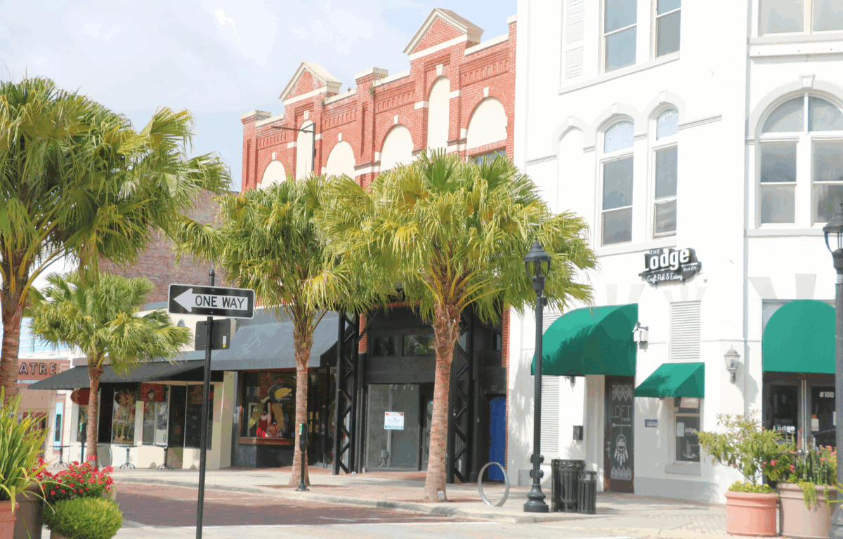 Historic brick buildings with a mix of red brick and white facades, with green awnings over storefronts. A sign for "The Lodge" bar and restaurant is visible on a white building to the right. A "One Way" street sign points left and colorful flower planters decorate the sidewalk.