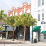 Historic brick buildings with a mix of red brick and white facades, with green awnings over storefronts. A sign for "The Lodge" bar and restaurant is visible on a white building to the right. A "One Way" street sign points left and colorful flower planters decorate the sidewalk.