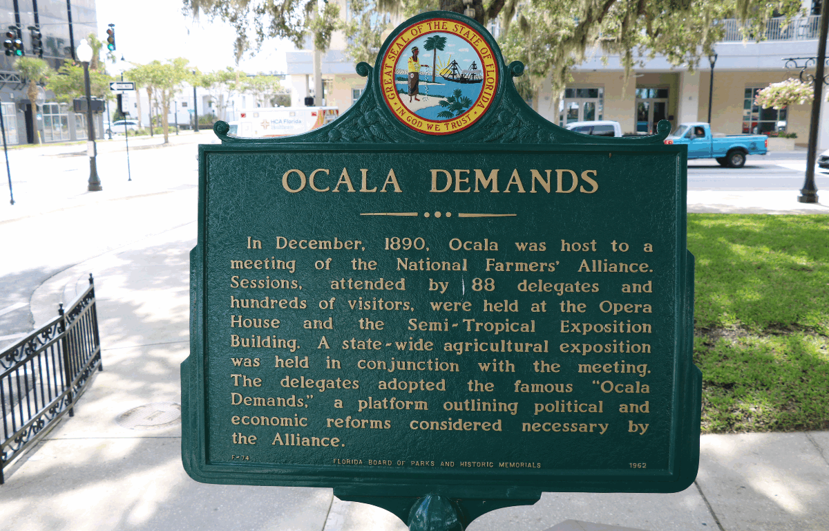 Dark green square sign that displays the Ocala Demands in beige with circular Ocala logo on top. In the background is bustling streets with cars passing by, a traffic light, a pole with a pot of pink flowers hanging from it, a beige building and roads leading in multiple directions.