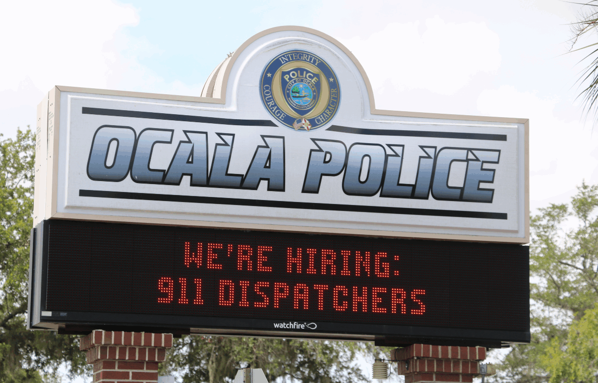 A sign for the Ocala Police Department with a digital message board below. The top of the sign displays the Ocala Police logo with the words "Integrity, Courage, Character" and large text reading "OCALA POLICE." The digital board underneath shows a scrolling message in red text that reads, "WE'RE HIRING: 911 DISPATCHERS." The sign is supported by red brick columns and surrounded by trees in the background.