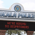 A sign for the Ocala Police Department with a digital message board below. The top of the sign displays the Ocala Police logo with the words "Integrity, Courage, Character" and large text reading "OCALA POLICE." The digital board underneath shows a scrolling message in red text that reads, "WE'RE HIRING: 911 DISPATCHERS." The sign is supported by red brick columns and surrounded by trees in the background.