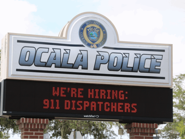 A sign for the Ocala Police Department with a digital message board below. The top of the sign displays the Ocala Police logo with the words "Integrity, Courage, Character" and large text reading "OCALA POLICE." The digital board underneath shows a scrolling message in red text that reads, "WE'RE HIRING: 911 DISPATCHERS." The sign is supported by red brick columns and surrounded by trees in the background.
