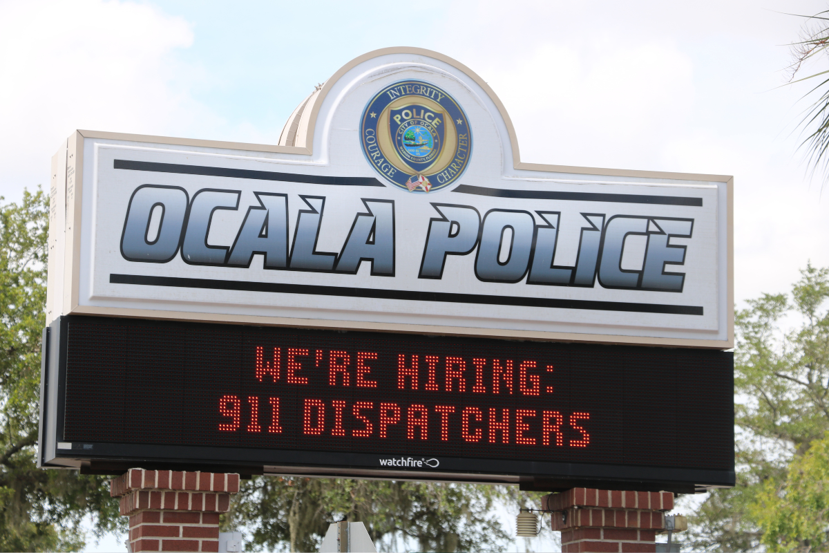 A sign for the Ocala Police Department with a digital message board below. The top of the sign displays the Ocala Police logo with the words "Integrity, Courage, Character" and large text reading "OCALA POLICE." The digital board underneath shows a scrolling message in red text that reads, "WE'RE HIRING: 911 DISPATCHERS." The sign is supported by red brick columns and surrounded by trees in the background.