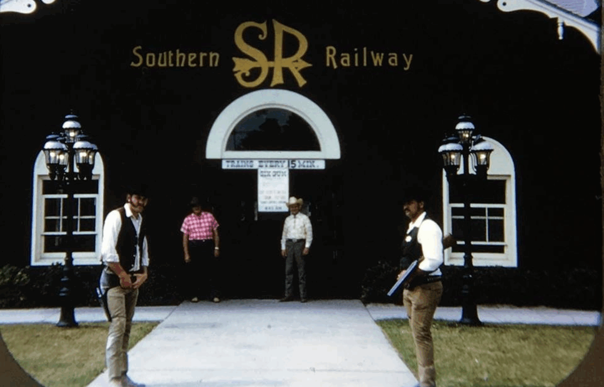 A black building with signage advertising the Southern Railway states "Trains every 15 minutes." Cowboys gather around the entrance of the station.