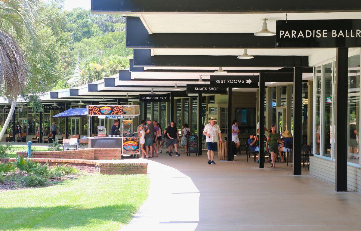 People walk and gather in a shaded outdoor shopping and dining area. Signs above indicate locations such as "Paradise Ballroom," "Rest Rooms," "Snack Shop," "Springside Café," and "Discovery Center." A food cart selling pretzels is positioned along the walkway, and some visitors are seated at tables near storefronts. The area is surrounded by green grass and trees.