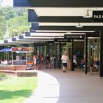People walk and gather in a shaded outdoor shopping and dining area. Signs above indicate locations such as "Paradise Ballroom," "Rest Rooms," "Snack Shop," "Springside Café," and "Discovery Center." A food cart selling pretzels is positioned along the walkway, and some visitors are seated at tables near storefronts. The area is surrounded by green grass and trees.