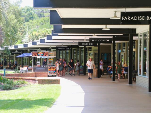 People walk and gather in a shaded outdoor shopping and dining area. Signs above indicate locations such as "Paradise Ballroom," "Rest Rooms," "Snack Shop," "Springside Café," and "Discovery Center." A food cart selling pretzels is positioned along the walkway, and some visitors are seated at tables near storefronts. The area is surrounded by green grass and trees.