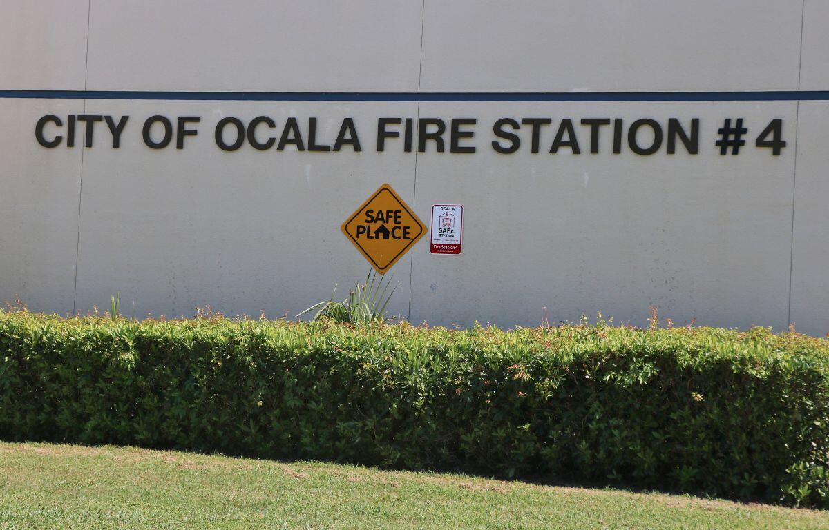 Extreme close-up of a beige building with green bushes in front of it and bold letters that read, "City of Ocala Fire Station #4." Toward the bottom of the letters is a yellow diamond-shaped sign that reads, "Safe Place." To the left of that sign is a red and white sign.