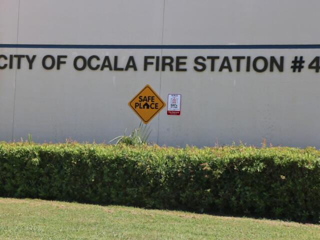 Extreme close-up of a beige building with green bushes in front of it and bold letters that read, "City of Ocala Fire Station #4." Toward the bottom of the letters is a yellow diamond-shaped sign that reads, "Safe Place." To the left of that sign is a red and white sign.