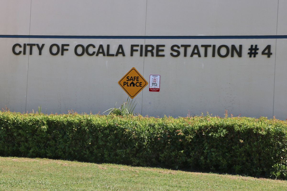 Extreme close-up of a beige building with green bushes in front of it and bold letters that read, "City of Ocala Fire Station #4." Toward the bottom of the letters is a yellow diamond-shaped sign that reads, "Safe Place." To the left of that sign is a red and white sign.