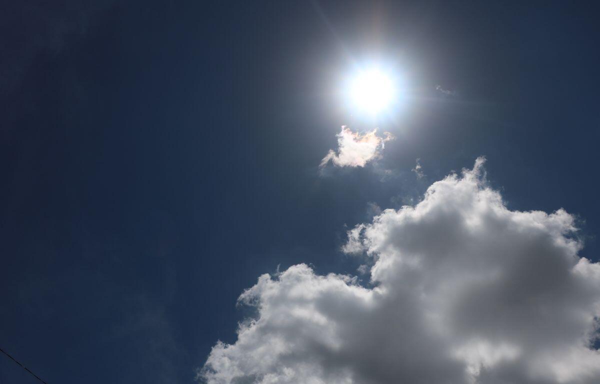 The sun shining brightly in a clear blue sky with a small cloud being brightened by the sun and a large white cloud below it.