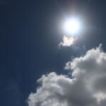 The sun shining brightly in a clear blue sky with a small cloud being brightened by the sun and a large white cloud below it.
