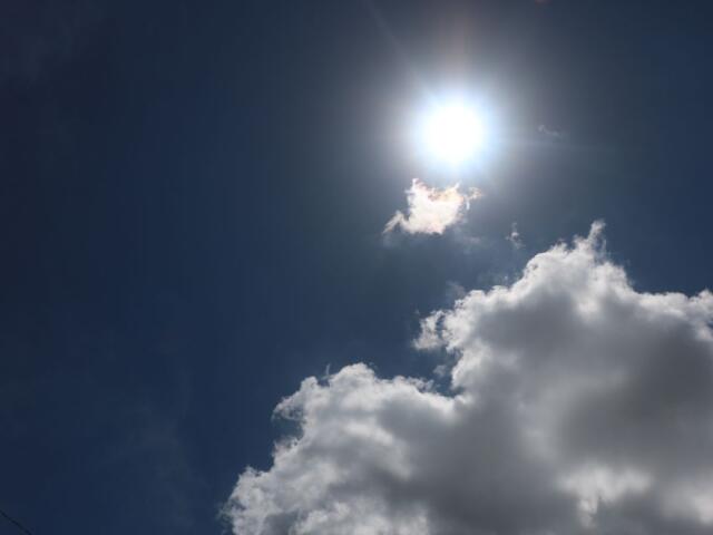 The sun shining brightly in a clear blue sky with a small cloud being brightened by the sun and a large white cloud below it.