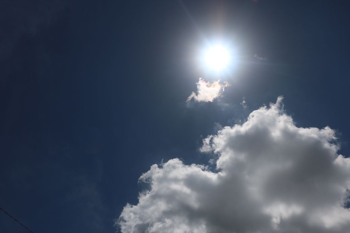 The sun shining brightly in a clear blue sky with a small cloud being brightened by the sun and a large white cloud below it.