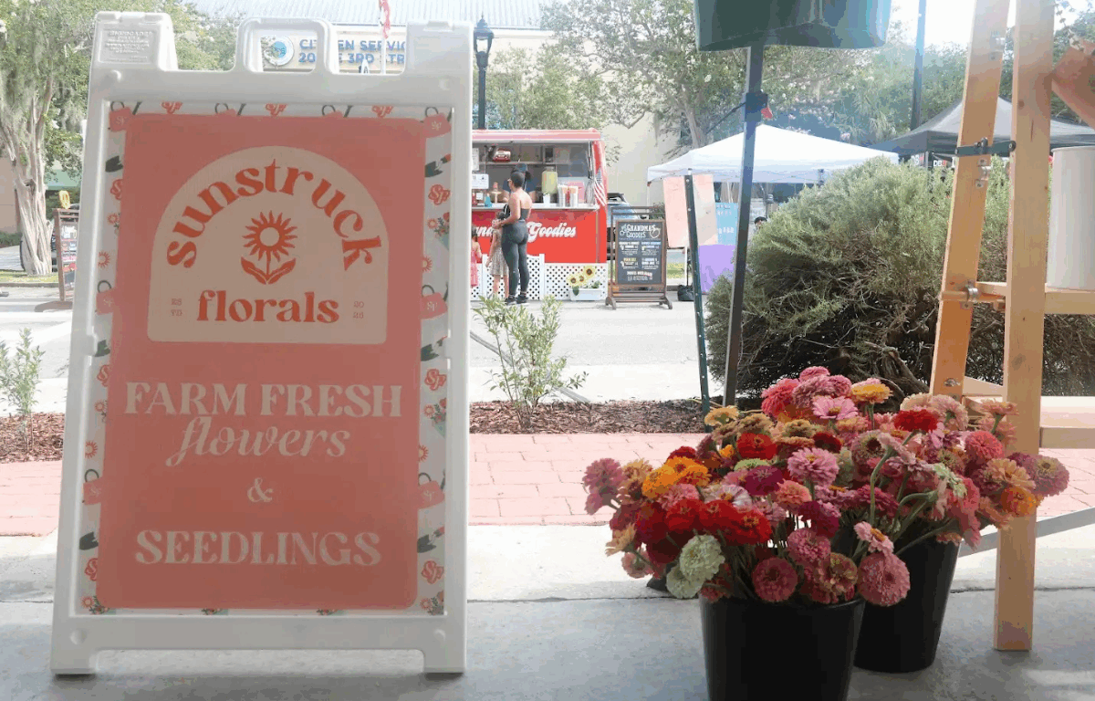 Promotional signage for the Sunstruck Florals stand advertises fresh flowers and seedlings with a view of marketgoers and other vendors in the background.