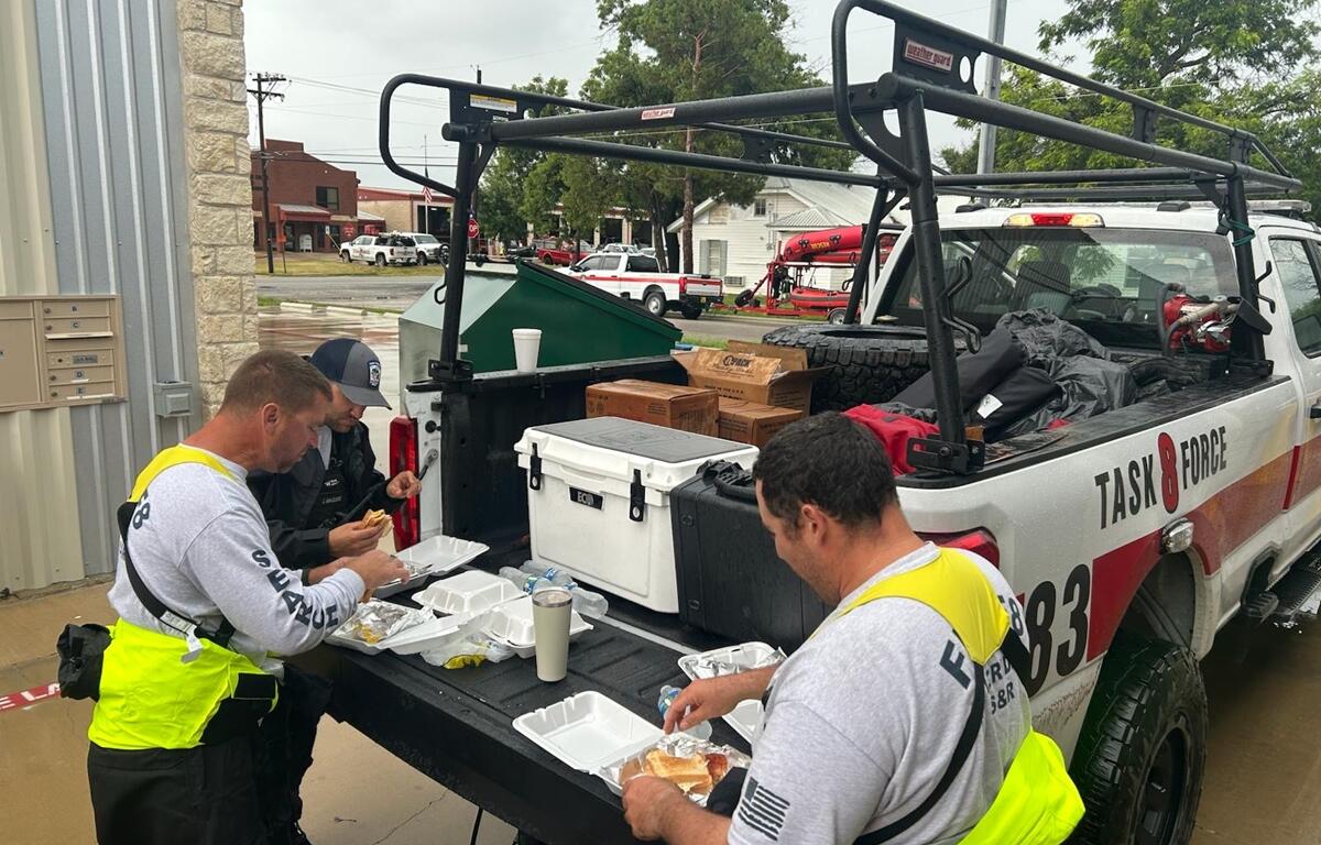 Three Task Force 8 members eat lunch from takeout containers on the tailgate of a truck during flood response in Texas.