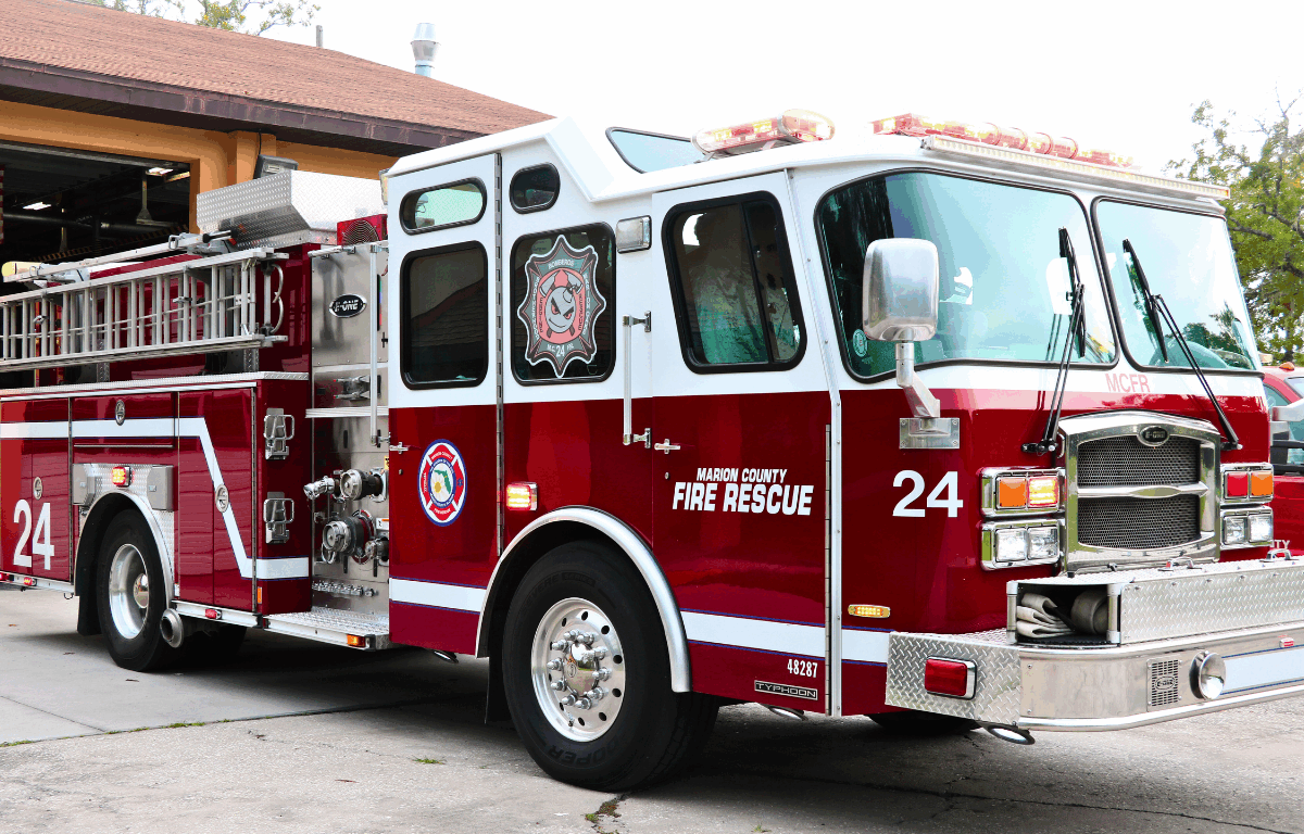 A red and white fire truck with "Marion County Fire Rescue" and "24" written on the side and a peach-colored fire station with an open garage door.