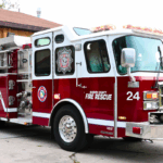 A red and white fire truck with "Marion County Fire Rescue" and "24" written on the side and a peach-colored fire station with an open garage door.