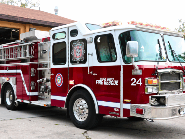 A red and white fire truck with "Marion County Fire Rescue" and "24" written on the side and a peach-colored fire station with an open garage door.