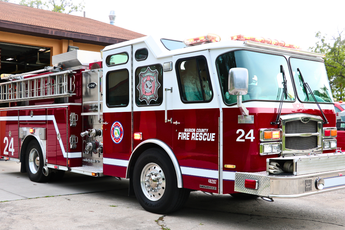 A red and white fire truck with "Marion County Fire Rescue" and "24" written on the side and a peach-colored fire station with an open garage door.