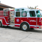A red and white fire truck with "Marion County Fire Rescue" and "24" written on the side and a peach-colored fire station with an open garage door.
