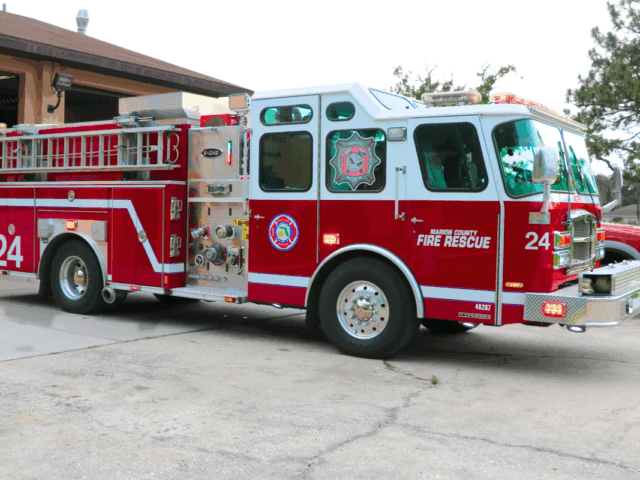 A red and white fire truck with "Marion County Fire Rescue" and "24" written on the side and a peach-colored fire station with an open garage door.