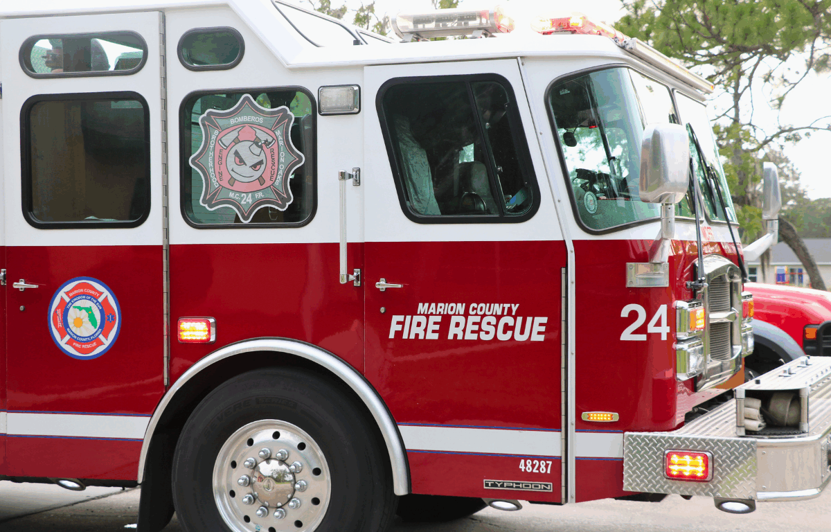 Close-up of a red fire truck with "Marion Country Fire Rescue" and "24" written on the side with a tree and light sky in the background.
