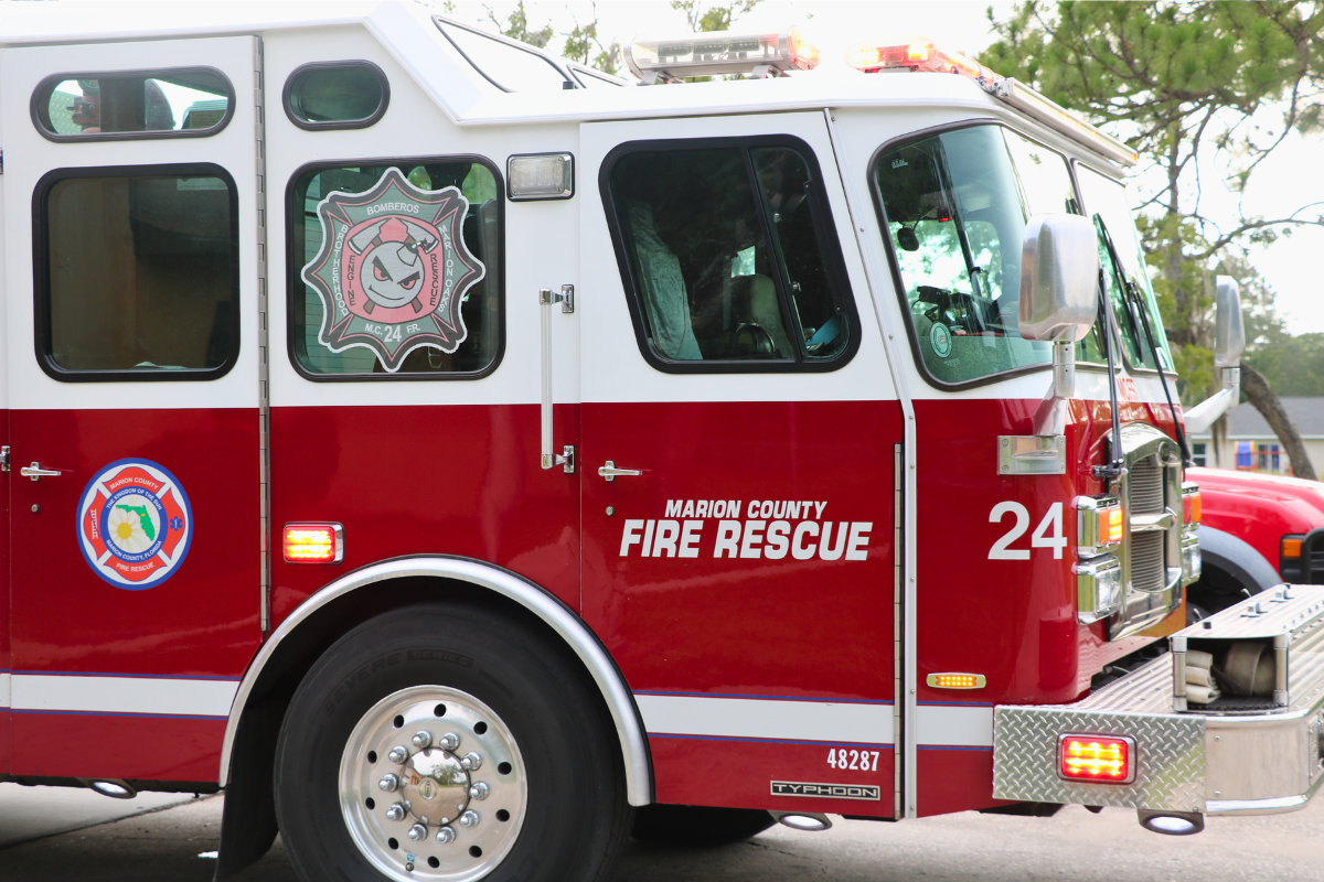 Close-up of a red fire truck with "Marion Country Fire Rescue" and "24" written on the side with a tree and light sky in the background.