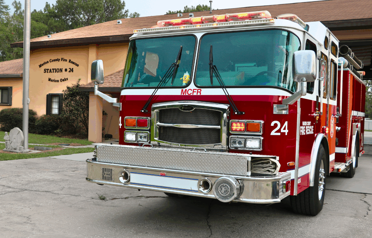 Front of a fire truck with "MCRF" written on the front with "24" and "Marion County Fire Rescue" on the side. A peach-colored fire station is in the background with "Marion County Fire Rescue Station # 24 Marion Oaks" written on the building to the left and flag pole to the left.