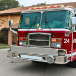 Front of a fire truck with "MCRF" written on the front with "24" and "Marion County Fire Rescue" on the side. A peach-colored fire station is in the background with "Marion County Fire Rescue Station # 24 Marion Oaks" written on the building to the left and flag pole to the left.