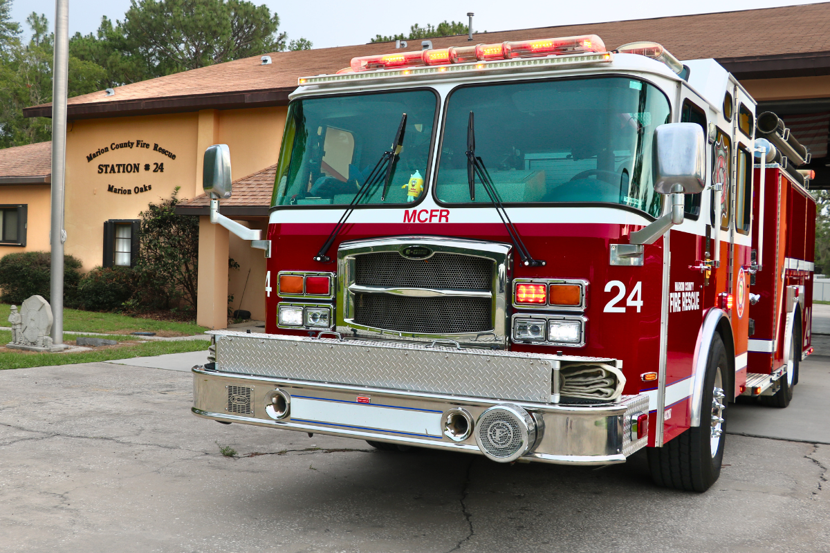 Front of a fire truck with "MCRF" written on the front with "24" and "Marion County Fire Rescue" on the side. A peach-colored fire station is in the background with "Marion County Fire Rescue Station # 24 Marion Oaks" written on the building to the left and flag pole to the left.