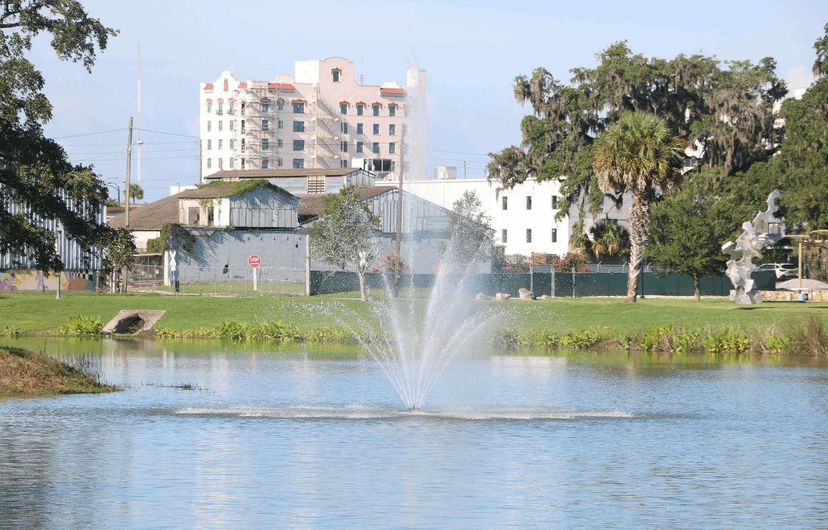 A body of water with a small fountain spouting water out in upward dispersal. Green grass and trees surround the water with a metal sculpture on the right and white and gray buildings directly behind the spouting water.